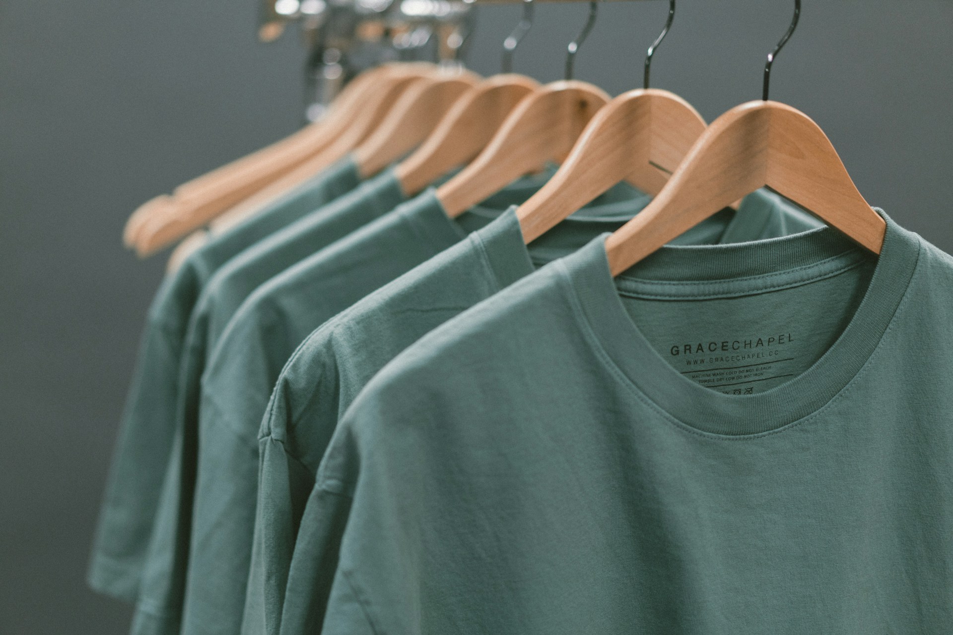 Dark seagreen t-Shirts on hangers in front of a grey background.
